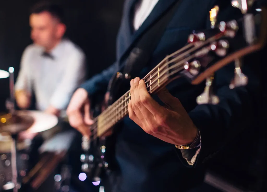 Close-up of hands playing bass guitar at an event, with the drummer in the background